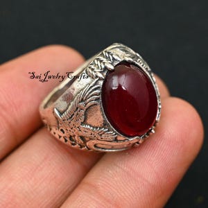May include: A silver ring featuring a deep red oval gemstone. The ring's band is decorated with dragon and floral patterns. The text "Sai Jwelry Crafts" is inscribed on the side of the ring.