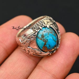 Puede incluir: Anillo de plata con una gran piedra turquesa ovalada con vetas marrones. El anillo tiene un diseño ornamentado con motivos de dragones o pájaros. El texto "Sai Jewelry Crafts" es visible en el anillo.