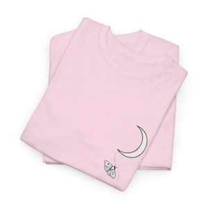 May include: Two pale pink t-shirts are stacked. The top one features a cream-coloured crescent moon and a black and white butterfly design. The butterfly appears to be flying away from the moon.