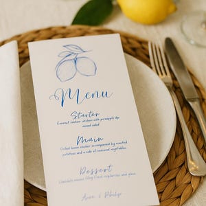 Peut inclure: Un menu avec une illustration de citron bleu et une écriture manuscrite. Le menu détaille une entrée, un plat principal et un dessert. La carte est posée sur une assiette blanche, avec des couverts et un dessous de plat tressé.