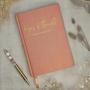 May include: A coral journal with gold lettering reading "Pages of Strength" and "walking through FMDS". A gold pen with a floral design and a cup of coffee are also in the image. Dried flowers are scattered around the journal.
