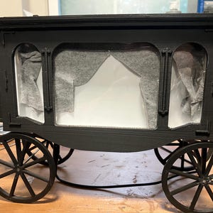 Puede incluir: Un modelo de carruaje de caballos impreso en 3D de color negro, con una cabina rectangular, aberturas de ventanas arqueadas, cortinas de tela gris drapeadas en el interior y cuatro ruedas de radios. La estructura tiene un techo plano.