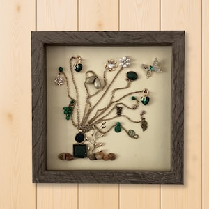 May include: A framed jewelry display featuring necklaces and earrings arranged to resemble a tree. The jewelry includes green and clear gemstones, gold-toned chains, and a butterfly brooch. The frame is a dark gray color.