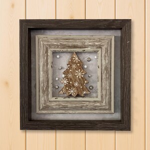 May include: A framed Christmas decoration featuring a wooden tree with snowflake cutouts. The tree is surrounded by small silver and white ornaments. The frame is a dark brown color, with a lighter, distressed frame inside.