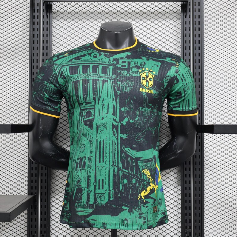 K&ouml;nnte beinhalten: Gr&uuml;nes und schwarzes Brasilien-Fu&szlig;balltrikot mit einem Stadtbild-Design. Das Trikot hat einen gelben Kragen und &Auml;rmelbesatz. Das Brasilien-Wappen und das Wort "BRASIL" befinden sich auf der Brust.