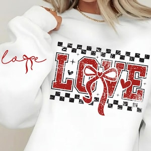 Pode incluir: Camisola branca com a palavra "LOVE" em letras vermelhas desgastadas, adornada com um laço e corações. O desenho é emoldurado por um padrão xadrez preto e branco. A palavra "love" também está escrita em vermelho na manga.
