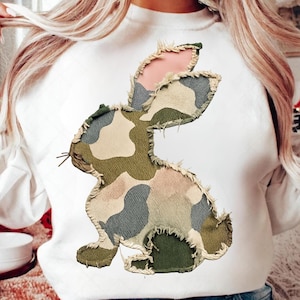 Op de afbeelding: Witte sweatshirt met een groot, geappliqueerd konijnontwerp. Het konijn is gemaakt van camouflage stof in groen, grijs en roze, met gerafelde randen. De sweatshirt heeft een ronde hals en lange mouwen.