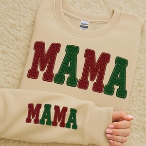 May include: Cream-colored sweatshirt with the word "MAMA" in a knitted design. The letters alternate between burgundy and green. The same design is repeated on the sleeve. A cozy and comfortable apparel item.