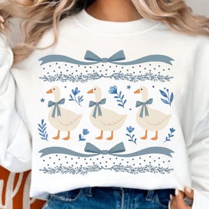 Op de afbeelding: Witte sweatshirt met een grillig ontwerp van drie ganzen met blauwe strikken, omringd door bloemen- en lintaccenten. Het ontwerp bevat blauwe bloemen en decoratieve elementen, wat een charmante en pastorale esthetiek creëert.