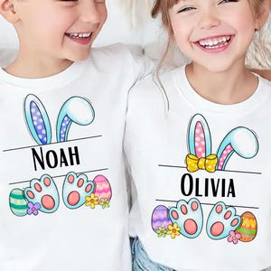 Op de afbeelding: Twee witte T-shirts met paasthema's. Elk shirt heeft konijnenoren, een naam, pootafdrukken en versierde paaseieren. Het ontwerp bevat pastelkleuren en bloemaccenten, perfect voor Pasen.