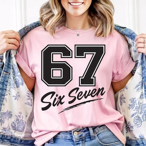 Könnte beinhalten: Ein hellrosa T-Shirt mit der Zahl "67" in schwarzer Schrift im College-Stil. Darunter stehen die Worte "Six Seven" in kursiver Schrift. Das T-Shirt ist Teil einer lässigen Bekleidungskollektion.