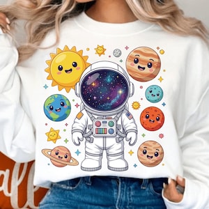 Puede incluir: Sudadera blanca con un astronauta de dibujos animados rodeado de planetas sonrientes, estrellas y un sol. El casco del astronauta muestra una galaxia colorida. El diseño incluye una Tierra sonriente, Saturno y otros planetas.