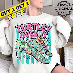 Op de afbeelding: Witte sweatshirt met een kleurrijke afbeelding van een zeeschildpad en de tekst "Turtley Over It". De schildpad heeft een regenboogschild, bloemen en sterren. Het sweatshirt heeft ook een "Koop 1 krijg 1 gratis" promotie.