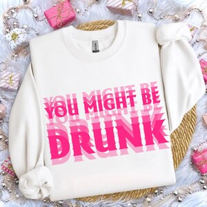 K&ouml;nnte beinhalten: Wei&szlig;es Sweatshirt mit dem Text "YOU MIGHT BE DRUNK" in &uuml;berlappenden rosa Buchstaben. Das Sweatshirt hat lange &Auml;rmel und einen Rundhalsausschnitt. Der Hintergrund umfasst Geschenkboxen und eine gewebte Matte.