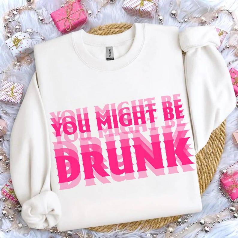 K&ouml;nnte beinhalten: Wei&szlig;es Sweatshirt mit dem Text "YOU MIGHT BE DRUNK" in &uuml;berlappenden rosa Buchstaben. Das Sweatshirt hat lange &Auml;rmel und einen Rundhalsausschnitt. Der Hintergrund umfasst Geschenkboxen und eine gewebte Matte.