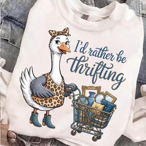 Könnte beinhalten: Weißes Sweatshirt mit einer Cartoon-Gans in einem Leopardenmuster-Outfit und blauen Stiefeln, die einen Einkaufswagen mit Vintage-Artikeln schiebt. Der Text "I'd rather be thrifting" ist in blauer Schrift geschrieben.