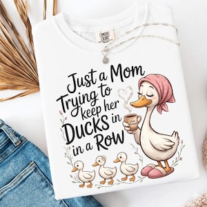 May include: White Comfort Colors sweatshirt with the text "Just a Mom Trying to Keep Her Ducks in a Row" and a cartoon duck design. The design features a duck wearing a pink headscarf and holding a coffee cup, with three ducklings.