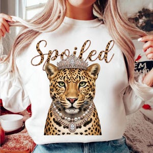 May include: White sweatshirt featuring a leopard wearing a crown and pearl necklace. The word "Spoiled" is written above the leopard in a leopard print font. The leopard has green eyes and a pink nose.