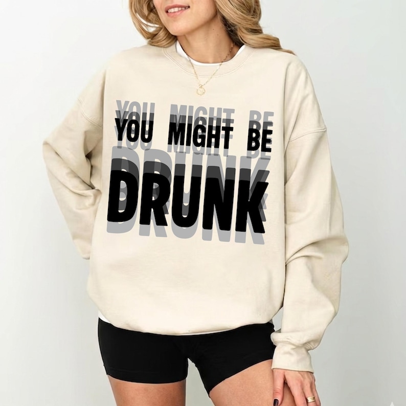 K&ouml;nnte beinhalten: Cremefarbenes Sweatshirt mit dem Text "YOU MIGHT BE DRUNK" in einem geschichteten schwarz-grauen Schriftzug. Das Sweatshirt ist ein l&auml;ssiger Stil, geeignet f&uuml;r den Alltag.