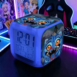 May include: A blue cube-shaped digital alarm clock with a cartoon cat design. The clock face displays the time, day, date, and temperature. The cat illustration features blue fur, orange eyes, and sharp teeth.