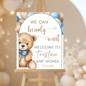 May include: A baby shower welcome sign on an easel. The sign features a teddy bear with a blue bow tie and the text "We can bearly wait" and "Welcome to Tristan Baby Shower 02.02.2026". The background includes blue and beige balloons.