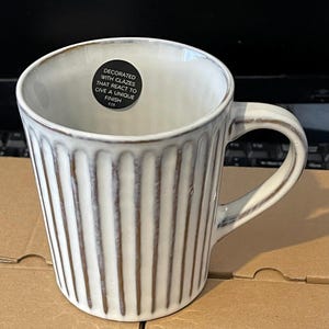 May include: White ceramic mug with vertical ribbed design and a brown rim. The mug has a curved handle and a sticker that reads "DECORATED WITH GLAZES THAT REACT TO GIVE A UNIQUE FINISH".