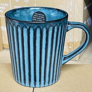 May include: A teal ceramic mug with vertical ribbed detailing and a curved handle. The mug has a glossy finish and a dark rim. A sticker on the mug reads "DECORATED WITH GLAZES THAT REACT TO GIVE A UNI..."