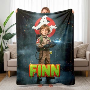 May include: A black blanket featuring a Ghostbusters theme. The blanket displays the Ghostbusters logo, a child dressed as a Ghostbuster, and the name "FINN" in green, dripping letters. The background has a dark, smoky effect.