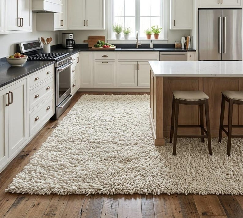 May include: A large, rectangular, off-white shag rug with a thick, high-pile texture covering the wooden floor in a kitchen. The rug features long, soft fibers that create a dense, plush surface, extending from the cabinets to the island.