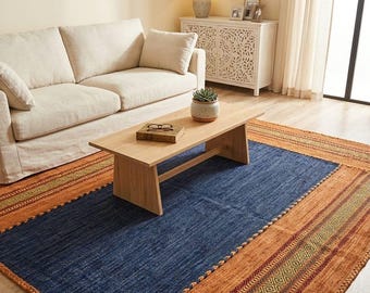 May include: A rectangular area rug featuring a large central navy blue panel bordered by wide bands of burnt orange. The orange borders are decorated with intricate geometric patterns in green and red, finished with a braided edge texture.