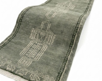 May include: A long, rectangular runner rug in a muted sage green color. The rug features a repeating geometric pattern in a lighter, off-white tone. It has a short, dense pile texture and is finished with a short, light-colored fringe at the ends.
