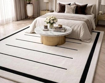 May include: A rectangular area rug featuring a cream-colored base with a thick black border and several horizontal black lines of varying lengths. The rug is positioned on a light wood floor in a bedroom setting, extending underneath a round coffee table.