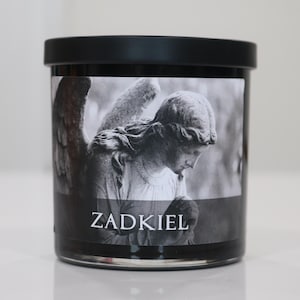 May include: A black candle jar with a black lid. The label features a grayscale image of an angel with wings, and the word "ZADKIEL" in white text.