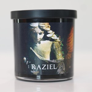May include: A black-lidded glass jar with a label featuring a statue of an angel. The angel is depicted in shades of gray and gold, with the word "RAZIEL" printed in white at the bottom of the label.