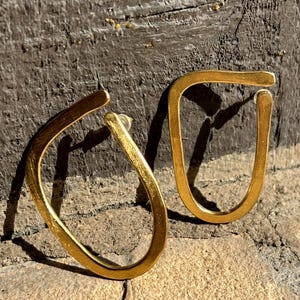 May include: A pair of gold-colored, open-hoop earrings. The earrings have a unique, irregular shape, with a slightly flattened, curved design. The earrings are displayed on a textured, neutral-toned surface, with a dark background.