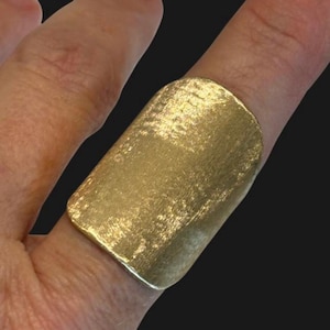 May include: A gold-colored ring with a rectangular face. The ring has a textured surface and is worn on a finger. The ring's design is simple and modern, with a brushed metal appearance.
