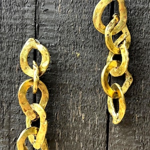 May include: Two gold-colored chain links hang vertically against a dark wood background. The links have an irregular, hammered texture, giving them a rustic appearance. The chains are connected in a series of three and four links.