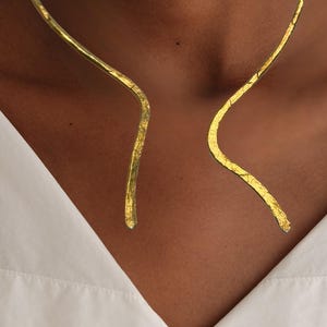 May include: A gold-colored necklace with a textured surface. The necklace curves around the neck, with a gap in the front. The person is wearing a white collared shirt.