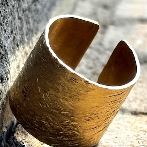 May include: A gold-colored cuff bracelet with a textured surface. The open-ended design allows for easy wear. The jewelry piece is set against a stone background, highlighting its metallic sheen.