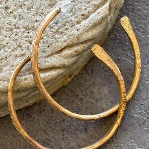 May include: A pair of gold-colored hoop earrings with a textured, hammered finish. The earrings are displayed on a light-colored stone surface, showcasing their warm metallic hue and unique design. The earrings have a post and backing.
