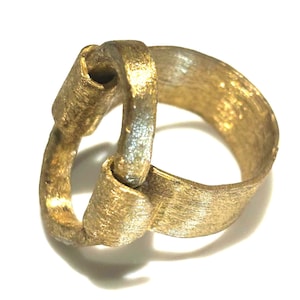 May include: A gold-colored ring with a unique interlocking design. The ring features a textured surface and a brushed metal appearance. The two interlocking loops create a continuous, flowing shape. The ring is likely made of metal.
