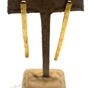May include: A pair of gold-colored, linear earrings. The earrings are displayed on a brown jewelry stand. The earrings have a slightly textured surface and a rectangular shape, with a rounded top. The earrings are hanging vertically.