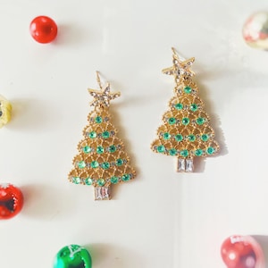 Puede incluir: Pendientes dorados con forma de árbol de Navidad con detalles de cristal verde y transparente. Cada pendiente tiene una estrella en la parte superior y un cristal rectangular en la base. Joyería festiva.