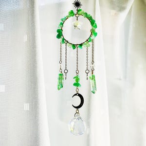 Puede incluir: Un atrapasoles decorativo con un marco circular adornado con piedras verdes y una estrella en la parte superior. Cadenas con cristales verdes, una luna creciente y una bola de cristal transparente cuelgan del marco. El atrapasoles cuelga frente a una cortina blanca.