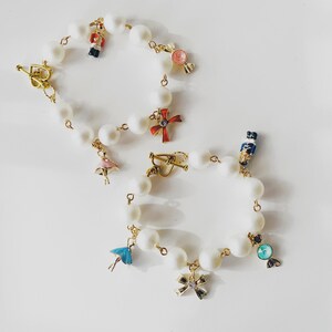 Pode incluir: Duas pulseiras de pérolas brancas com fechos dourados e amuletos decorativos. Os amuletos incluem uma bailarina, um quebra-nozes, um laço, uma cruz e um doce. As pulseiras estão sobre uma superfície branca.