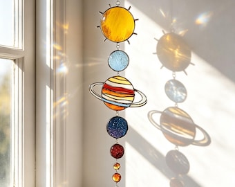 May include: A vertical stained glass suncatcher featuring a yellow sun at the top, followed by a light blue sphere, a large ringed planet with orange and white bands, a dark blue speckled sphere, a red sphere, and three smaller amber and blue spheres.