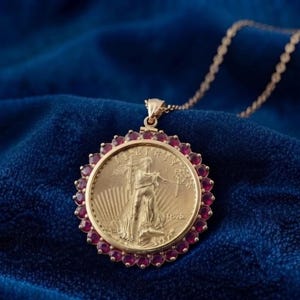 Op de afbeelding: Een gouden munt hanger ketting met een centrale gouden munt die een staande figuur met lichtstralen toont, omringd door een cirkelvormige rand van kleine, ronde, donkerrode edelstenen. De hanger hangt aan een fijne gouden schakelketting.