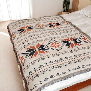 May include: A beige woven blanket with a geometric pattern in black, orange, and white. The blanket features a repeating design of star-like shapes and diamond patterns. The edges are finished with a fringe.