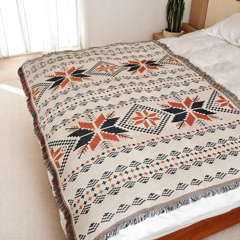 May include: A beige woven blanket with a geometric pattern in black, orange, and white. The blanket features a repeating design of star-like shapes and diamond patterns. The edges are finished with a fringe.