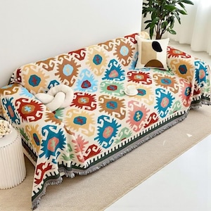 May include: A patterned sofa throw in a vibrant geometric design featuring red, blue, yellow, and green hues. The throw has a fringed border and is draped over a sofa. A decorative cushion and a white knot pillow are on the sofa.
